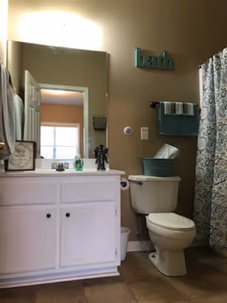 A bathroom with a white vanity cabinet and sink on the left, a large mirror above the sink, and a toilet next to it. There is a teal-colored wall decoration spelling 'bath' above the toilet, a teal towel hanging on a rack, and a teal basket on top of the toilet tank. A patterned shower curtain is visible on the right side.