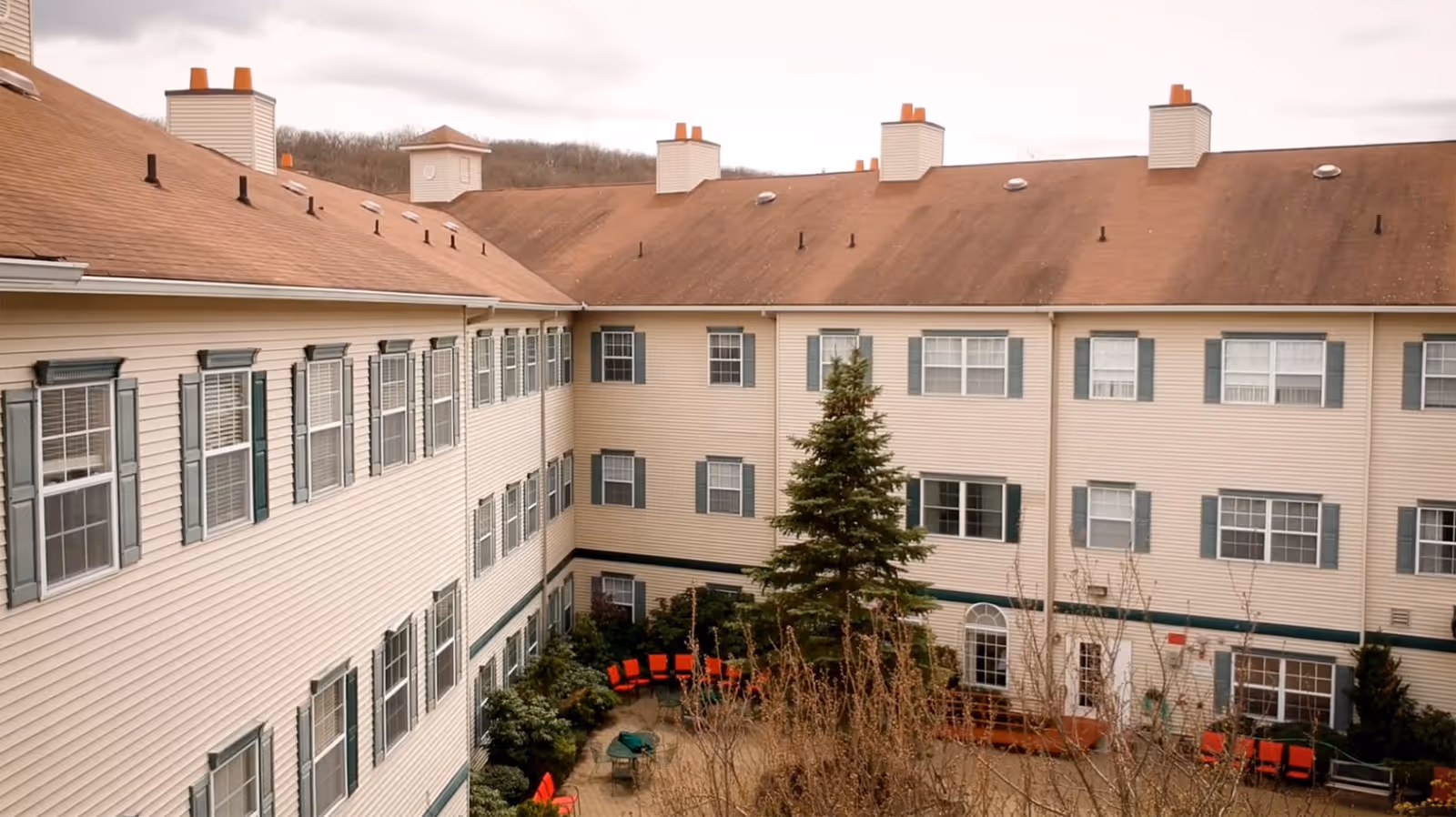 View of an outdoor courtyard area surrounded by a three-story beige building with green window shutters and a brown roof. The courtyard features a large evergreen tree, red chairs arranged in a semi-circle, and some shrubs and plants.
