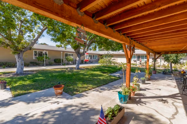 Covered wooden patio overlooking a grassy courtyard with potted plants, trees, and single-story care center buildings.