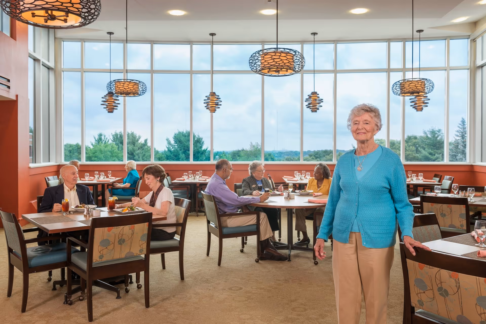 A bright dining room in a retirement community with large windows showing a view of trees outside. Several elderly people are seated at tables enjoying their meals, while one elderly woman in a blue cardigan stands smiling near the foreground. The room features modern hanging light fixtures and comfortable chairs with patterned upholstery.