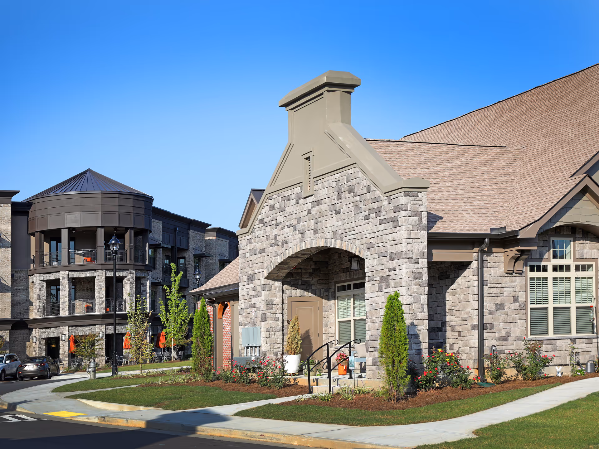 Exterior view of a senior living facility with stone and brick architecture, featuring a prominent chimney structure, landscaped garden with shrubs and flowers, and a multi-story building with balconies and outdoor seating in the background under a clear blue sky.