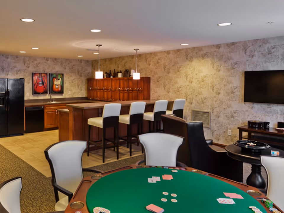 Communal interior room with a poker table in the foreground, a bar with high stools and kitchenette in the background, and a wall-mounted TV.