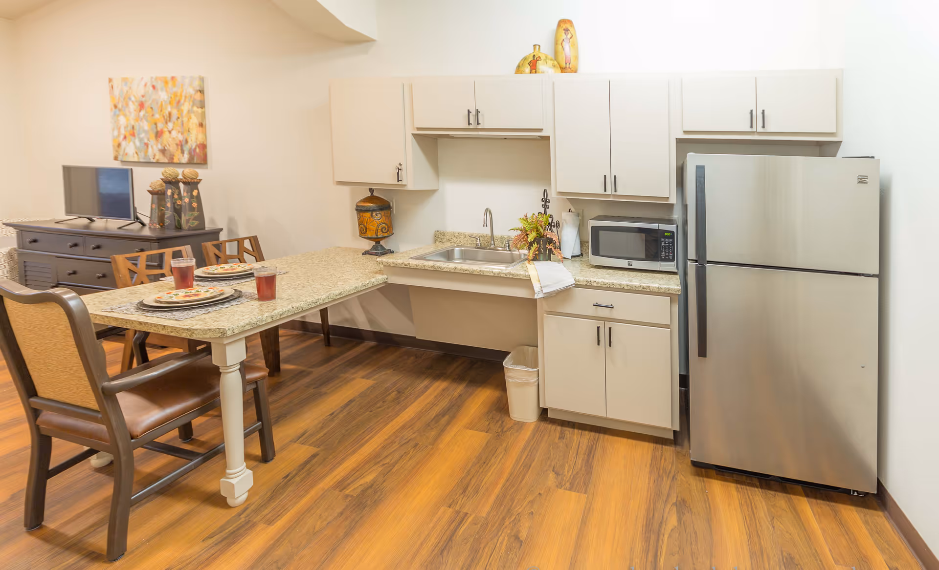 A small kitchen area with white cabinets, a stainless steel refrigerator, a microwave, and a sink. There is a granite countertop extending into a dining table with two place settings and two glasses of red beverage. In the background, there is a dark wooden dresser with a TV on top and a colorful abstract painting on the wall. The floor is wood with a warm tone.