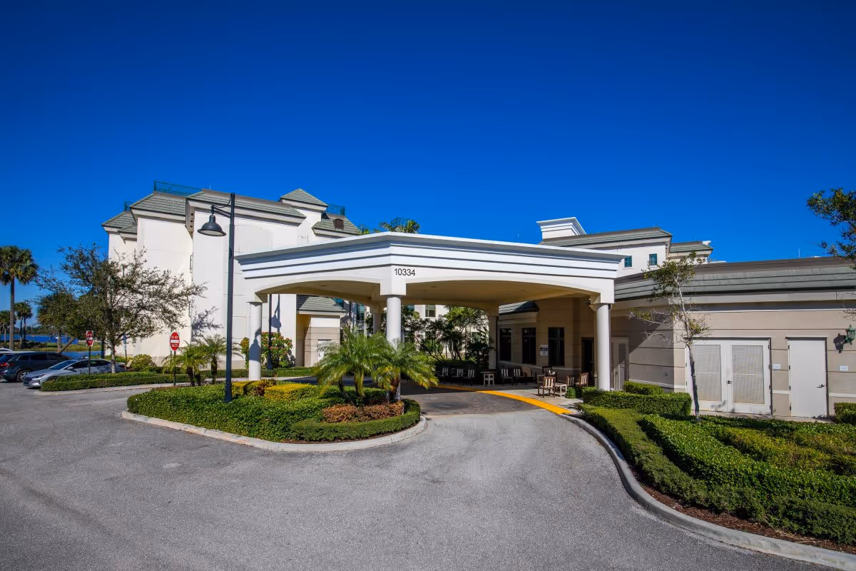 Front entrance of a senior living building with a covered porte-cochere, driveway and landscaped grounds under a clear blue sky.