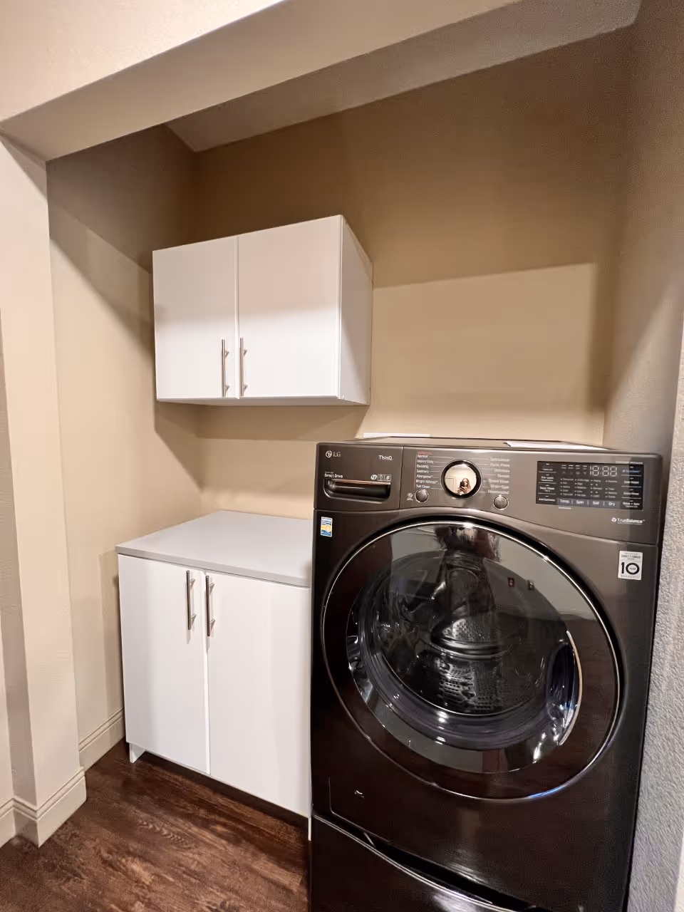 A laundry area with a black front-loading LG washing machine next to white cabinets, including a floor cabinet and a wall-mounted cabinet, with beige walls and dark wood flooring.