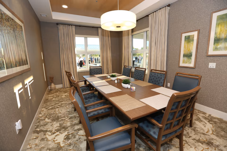 A well-lit dining room with a long wooden table set with placemats and small decorative plants. The room has eight cushioned chairs with wooden frames around the table. There are large windows with beige curtains letting in natural light, and framed artwork on the walls. A modern round ceiling light hangs above the table.