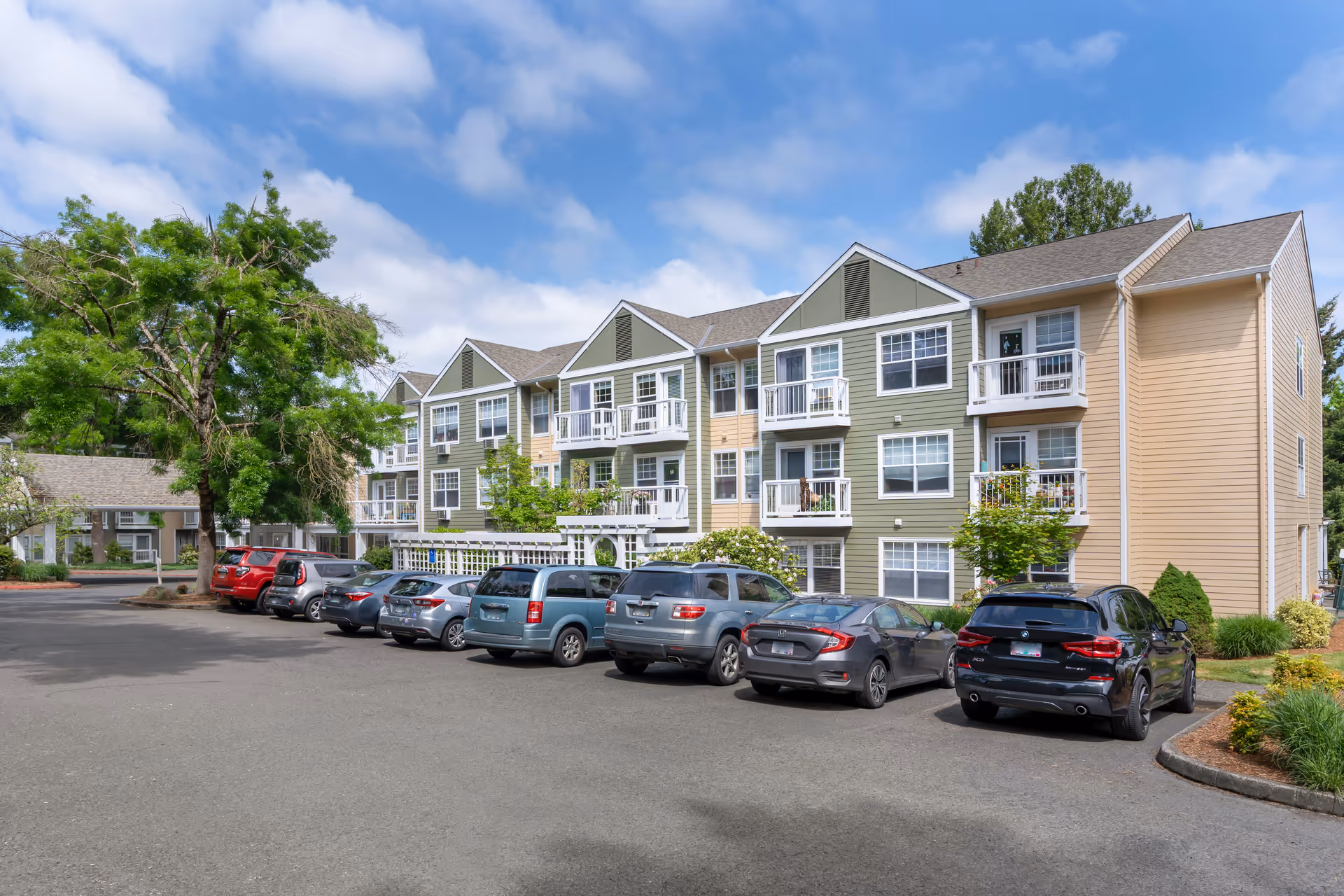 Exterior view of a multi-story residential building with beige and green siding, white balconies, and multiple windows. Several cars are parked in front of the building along a paved parking lot. There are trees and landscaped greenery around the building under a partly cloudy blue sky.