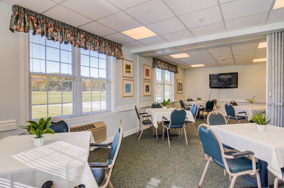 A bright dining room in a senior living community with several tables covered in white tablecloths, each adorned with a small green potted plant. Blue cushioned chairs surround the tables. Large windows with floral valances let in natural light, and framed artwork hangs on the walls. A flat-screen TV is mounted on the far wall.