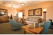 A cozy living room area with a striped sofa, two blue armchairs, a wooden coffee table with books on it, a wooden side table with a lamp, a wooden cabinet with a mirror above it, and framed artwork on the wall. The room is softly lit by ceiling lights and a floor lamp.