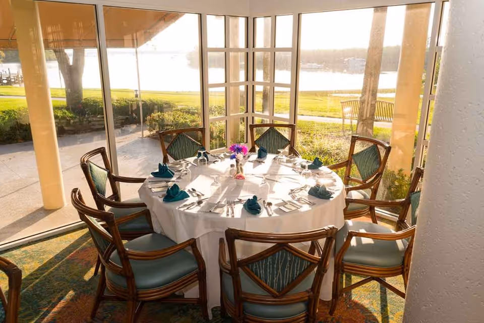 A round dining table set for eight people with white tablecloth, teal napkins, silverware, and glassware in a sunlit room with large windows overlooking a green lawn and water view outside.