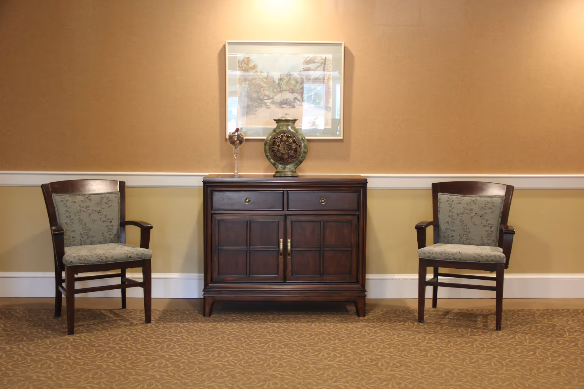 A simple interior setting featuring two wooden chairs with patterned cushions placed on either side of a dark wooden cabinet. On top of the cabinet is a decorative green vase and a glass container with some items inside. Above the cabinet hangs a framed artwork depicting a nature scene.
