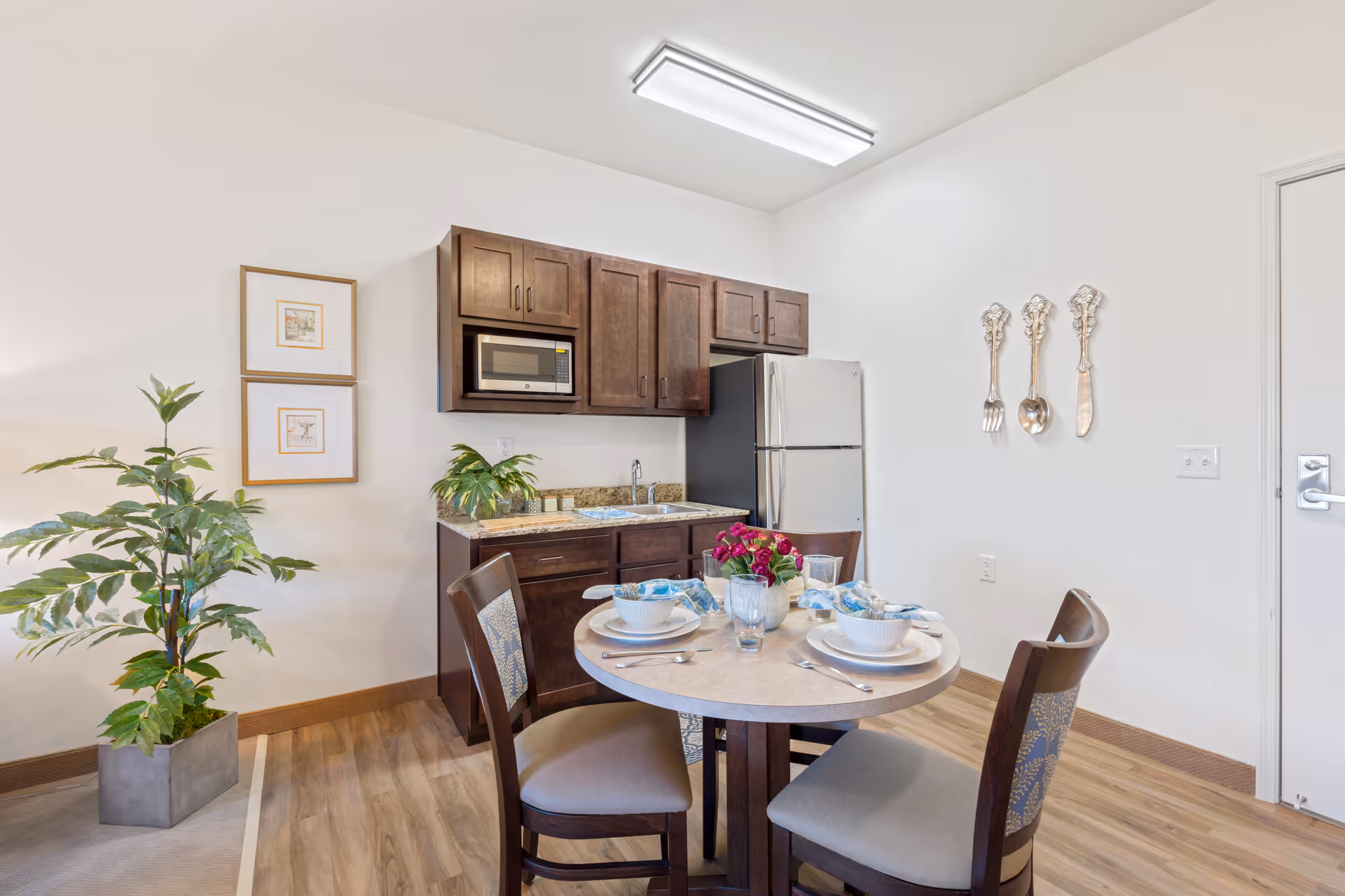 A small kitchen and dining area featuring dark wood cabinets, a microwave, a refrigerator, and a sink. In front of the kitchen is a round dining table set for four with white dishes, silverware, and a centerpiece of red flowers. The room has light-colored walls, wood flooring, a potted plant, two framed pictures on the wall, and decorative oversized silver utensils hanging on another wall.