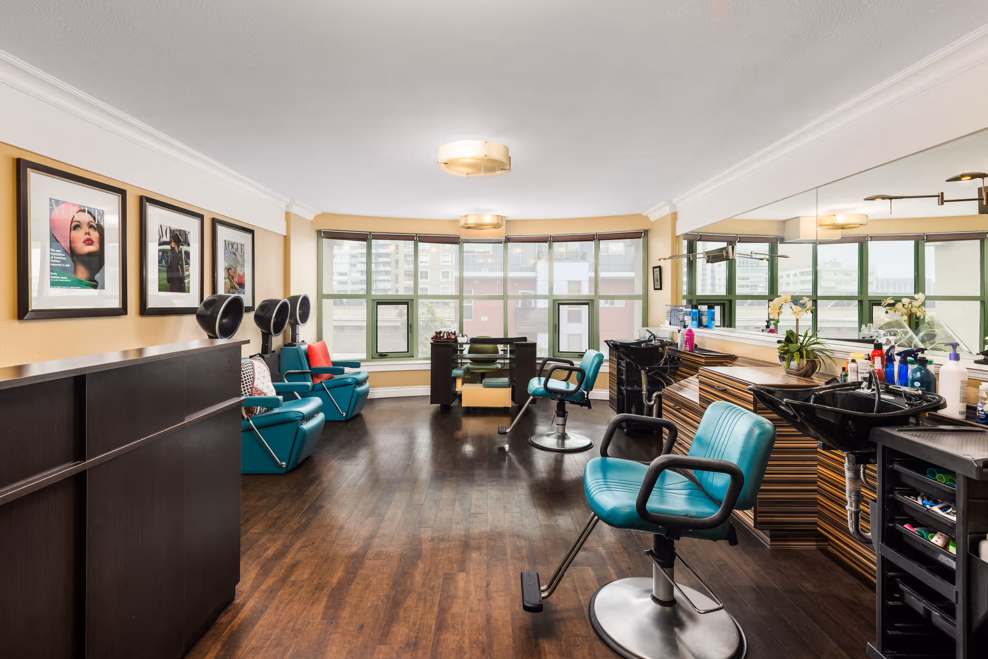 Interior view of a hair salon area with teal salon chairs, hair washing stations, a large mirror on the right wall, and framed pictures on the left wall. The room has large windows at the far end letting in natural light and dark wooden flooring.