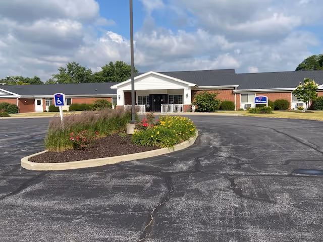 Front exterior view of Betz Nursing Home building with a circular driveway, landscaped island with flowers and shrubs, a handicap parking sign, and a covered entrance under a partly cloudy sky.