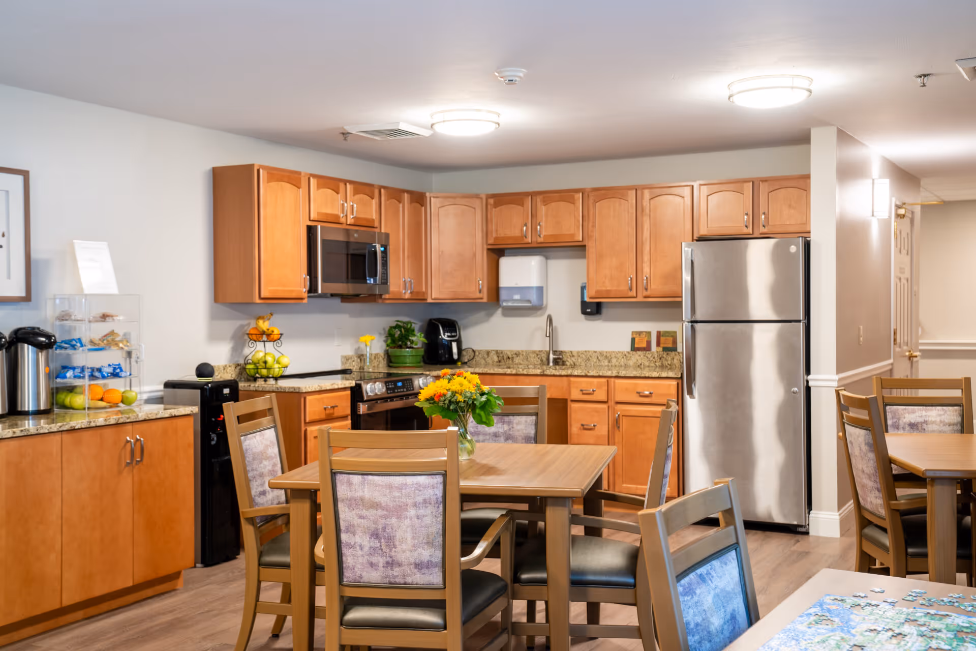 A well-lit kitchen and dining area in a senior living facility featuring wooden cabinets, a stainless steel refrigerator, microwave, stove, and a sink. There are multiple wooden dining tables with chairs, one table has a vase with yellow flowers. The countertops have a coffee maker, fruit, and snacks. The flooring is wood, and the walls are painted light gray.