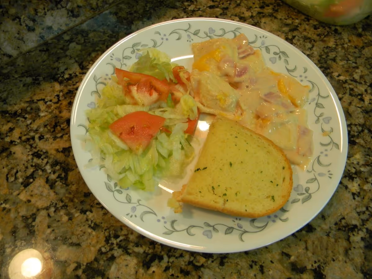 A plate with a serving of salad containing lettuce and tomato slices, a portion of ravioli with creamy sauce, and a slice of garlic bread on a granite countertop.