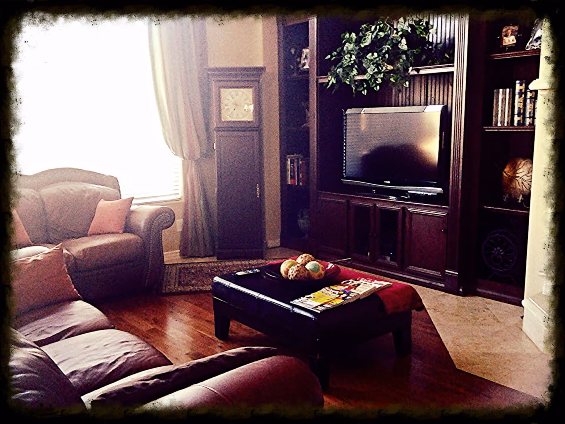 Cozy living room with leather sofas, a large wooden entertainment center with a flat-screen TV, a grandfather clock near a window with curtains, and a coffee table with decorative items and magazines.