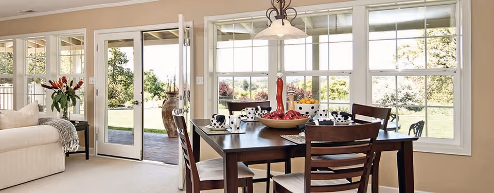 Bright open dining area with a dark wooden table and chairs beside large windows and a glass door opening to a garden, with a light-colored sofa visible at left.