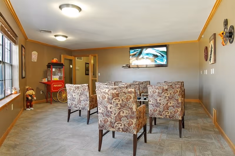 A cozy common area with patterned armchairs arranged in rows facing a wall-mounted flat screen TV. The room has beige walls with wooden trim, a popcorn machine in the corner, and decorative items including a scarecrow figure and wall art. Large windows on the left side let in natural light.