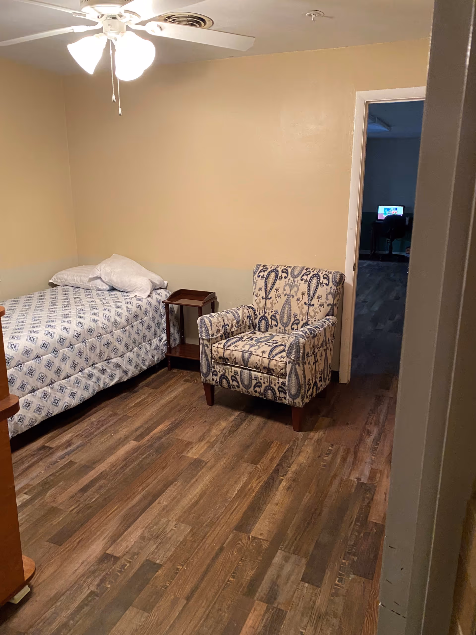 A small bedroom with a single bed covered in a patterned bedspread, a wooden side table, and an upholstered armchair with a paisley design. The room has wood flooring, beige walls, and a ceiling fan with lights. An open doorway shows a glimpse of another room with a desk and computer.