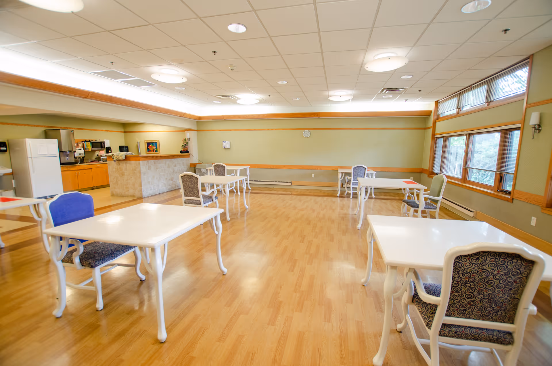 A spacious dining room with several white tables and cushioned chairs arranged with ample space between them. The room has light wood flooring, large windows with blinds on the right side, and a kitchenette area with a refrigerator, microwave, and coffee machine in the back left corner. The walls are painted light green with wooden trim, and the ceiling has recessed lighting and circular light fixtures.