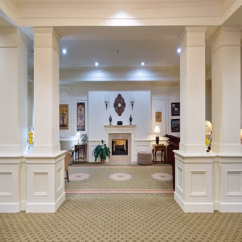 A spacious and elegant common area with beige carpet and white paneled columns. The room features a fireplace centered on the far wall, flanked by decorative wall sconces and vases. There are framed artworks on the walls, a piano on the right side, a small table with a lamp, and a plant near the fireplace. The lighting is bright with recessed ceiling lights.