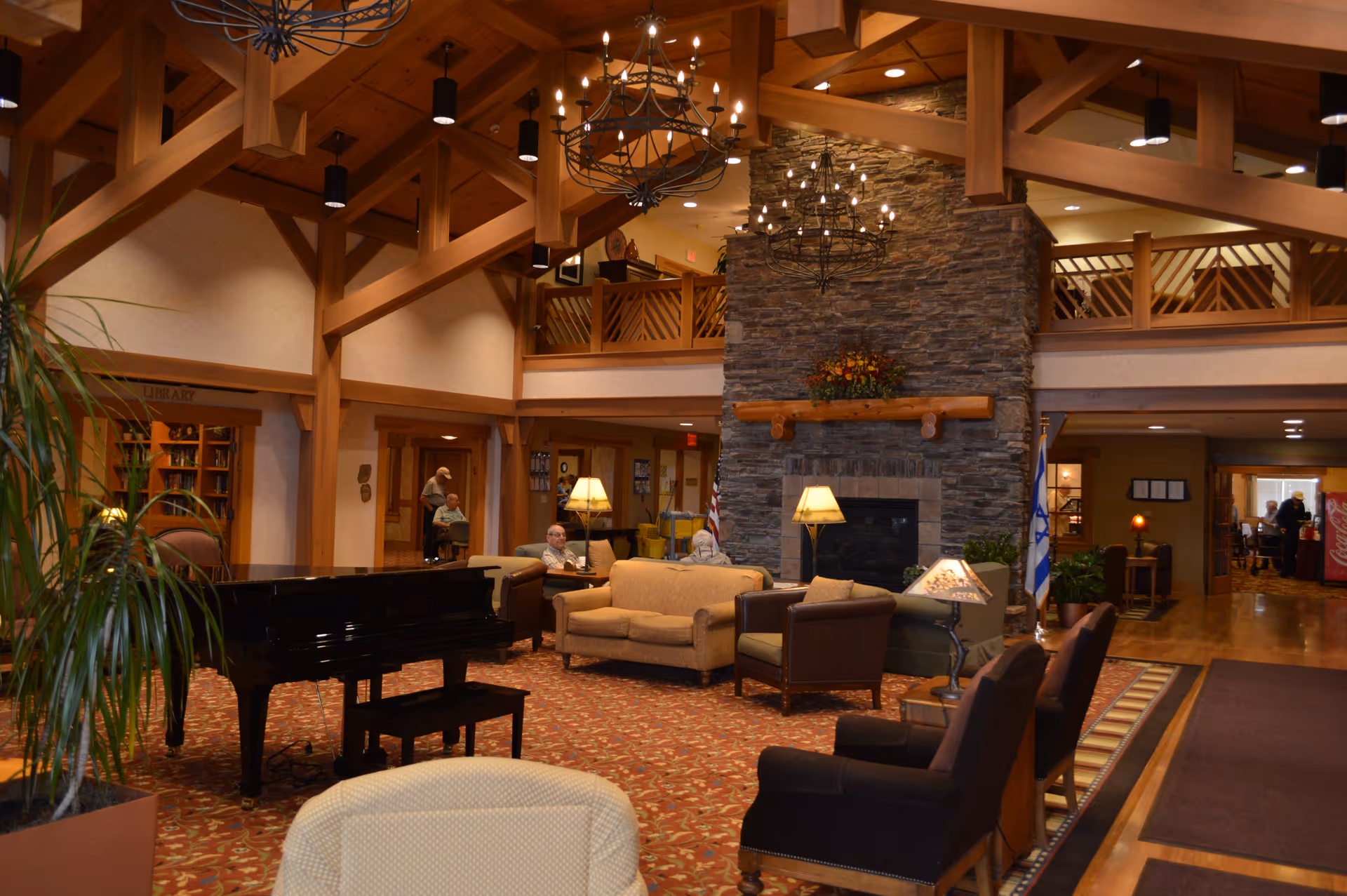A spacious senior living facility common area with wooden beams and a high ceiling. The room features a stone fireplace decorated with flowers, multiple seating arrangements including sofas and armchairs, a black grand piano, and warm lighting from chandeliers and lamps. There are a few elderly people seated and conversing, a library area visible on the left, and a hallway leading to other rooms in the background.