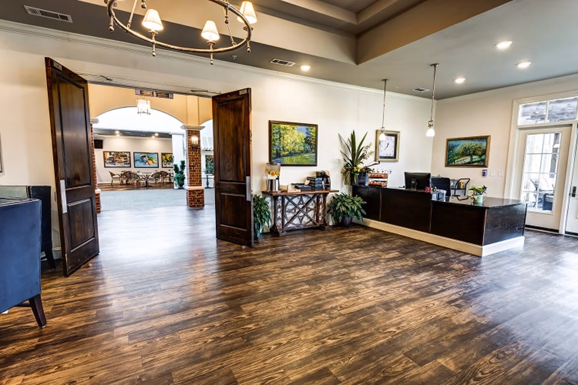 Interior view of a senior living facility reception area with wooden flooring, a dark wooden reception desk with a computer, potted plants, framed paintings on the walls, and double wooden doors leading to a common area with tables and chairs.
