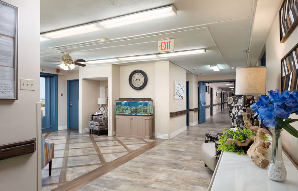 Well-lit interior lobby and hallway of a senior living facility featuring seating, an aquarium beneath a wall clock, decorative flowers, and doors leading to corridors.
