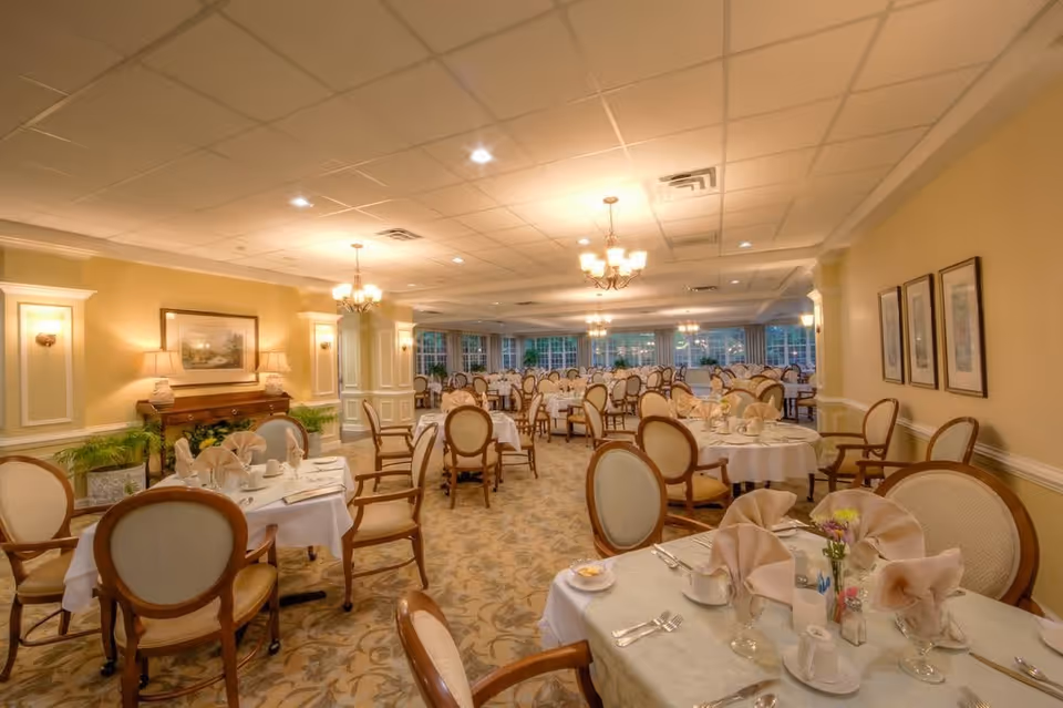 A spacious, elegantly decorated dining room with multiple round tables covered in white tablecloths, each set with folded napkins, cups, glasses, and silverware. The room features beige walls, framed artwork, chandeliers, and large windows along one side allowing natural light to enter.
