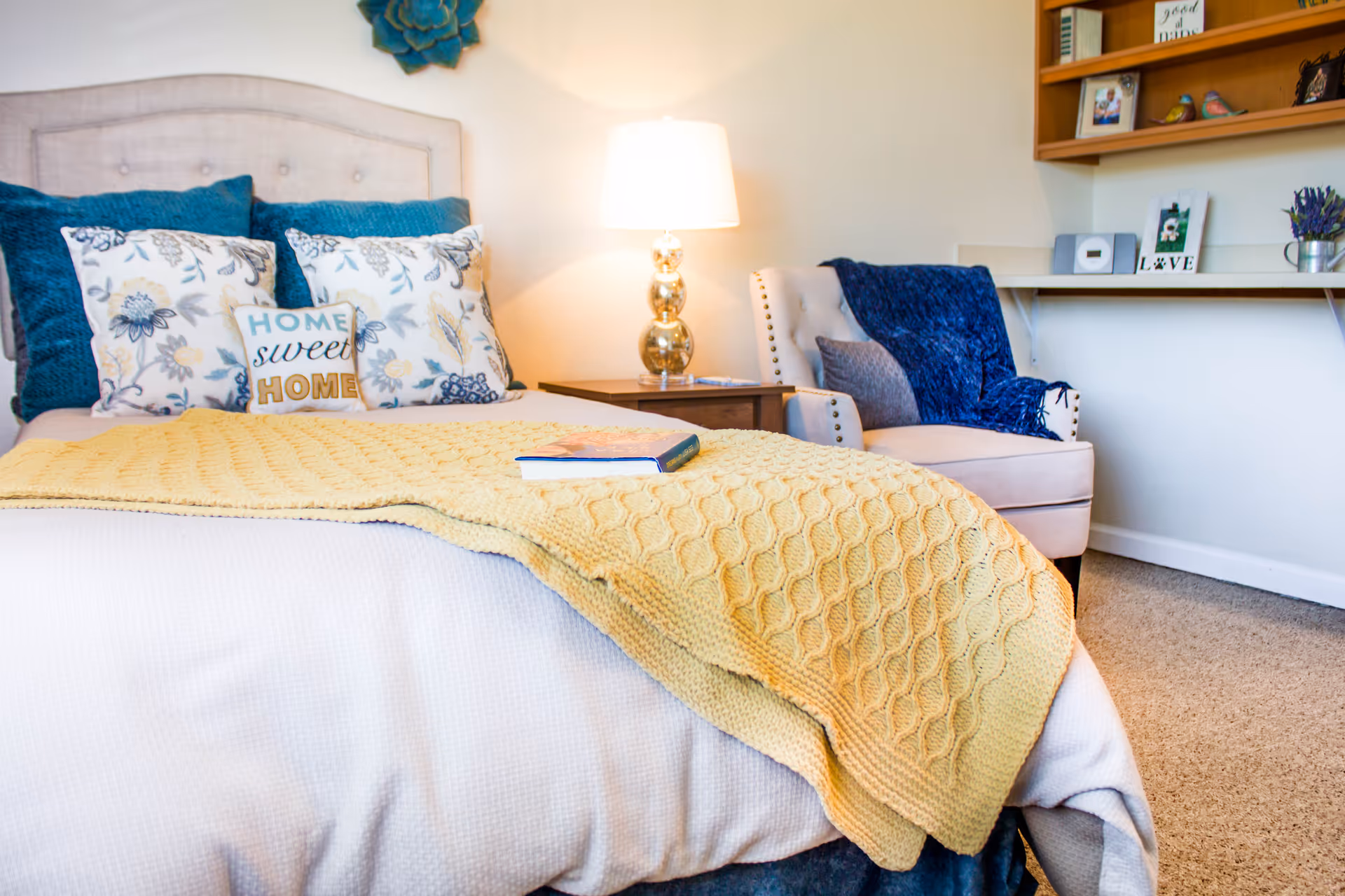 A cozy bedroom with a neatly made bed featuring floral and blue pillows, a small pillow with the words 'HOME sweet HOME', and a yellow knitted blanket. Next to the bed is a wooden nightstand with a modern lamp. There is a beige armchair with a blue throw blanket and a small pillow, and a wall-mounted shelf with decorative items and books.