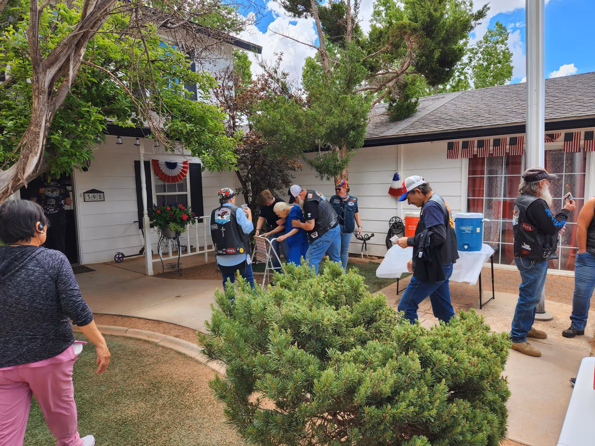 A group of visitors, including people in biker vests, assisting a resident with a walker outside the front entrance of a small senior living building decorated with American flags.