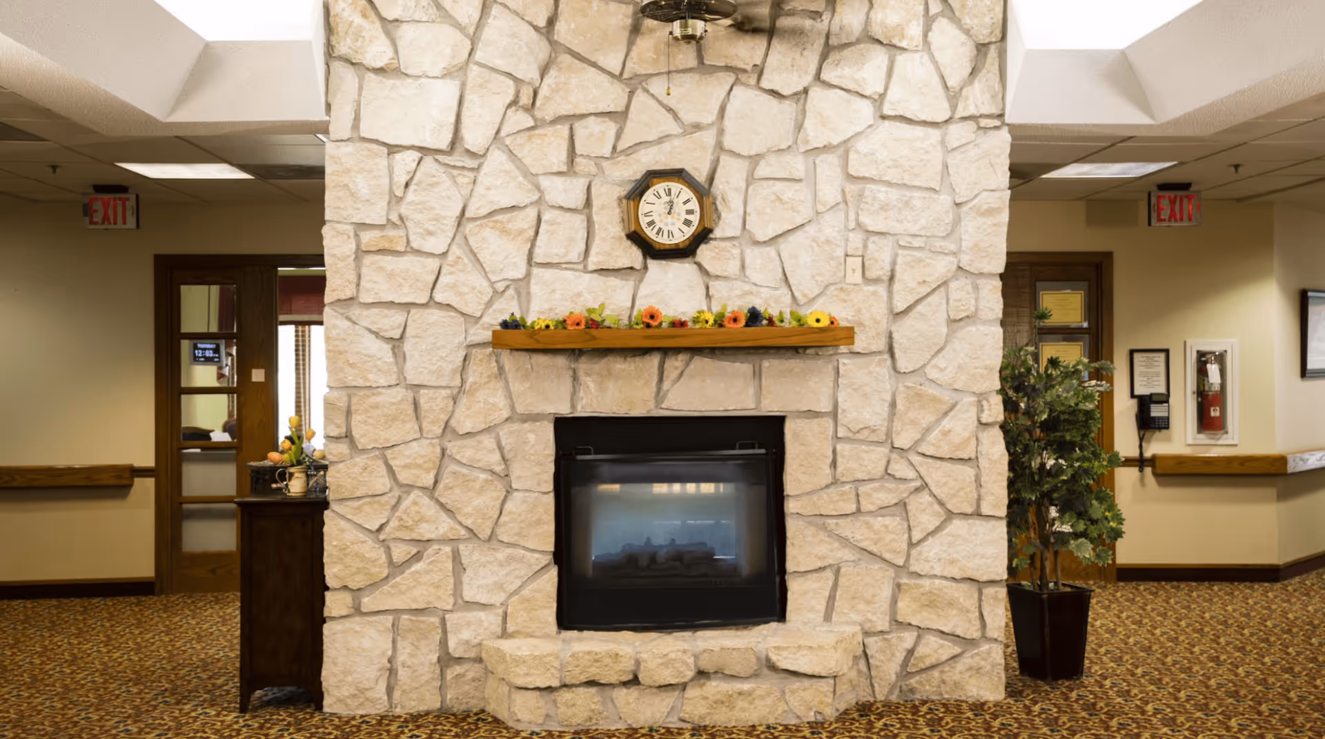 Stone fireplace with a mantel and clock in a carpeted assisted living facility common area with doors and exit signs.