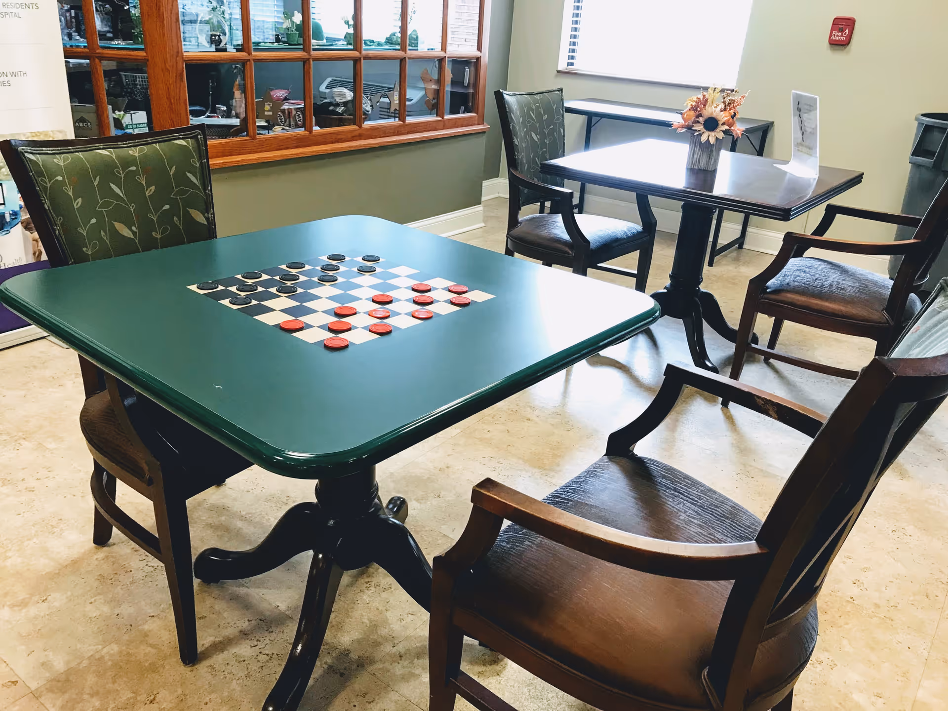 A room with two tables and several chairs. The table in the foreground has a built-in checkerboard with red and black checkers arranged for a game. The table in the background has a small flower arrangement and a sign. The room has a window with wooden framing and a fire alarm on the wall.