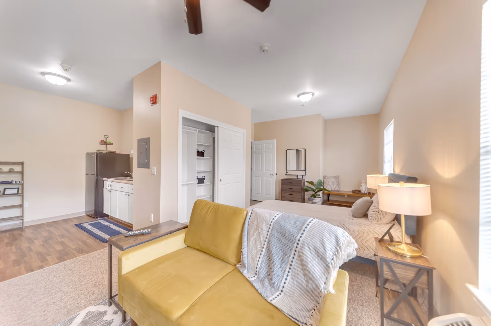Open studio living space with a yellow sofa in the foreground, a bed, kitchenette and dresser in a softly lit room.