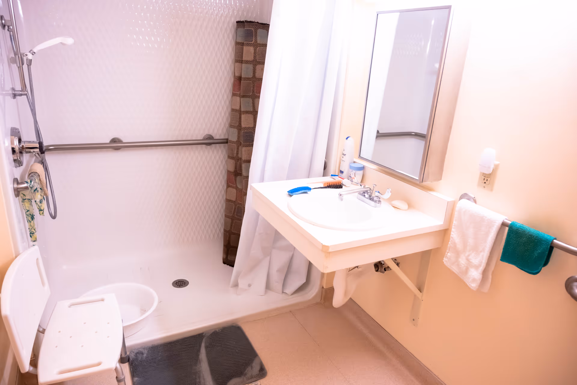 A clean bathroom with a walk-in shower featuring a handheld showerhead, a white shower chair, and a grab bar. There is a white sink with a mirror above it, a soap bar, and various toiletries on the sink counter. Two towels, one white and one green, hang on a towel rack next to the sink.