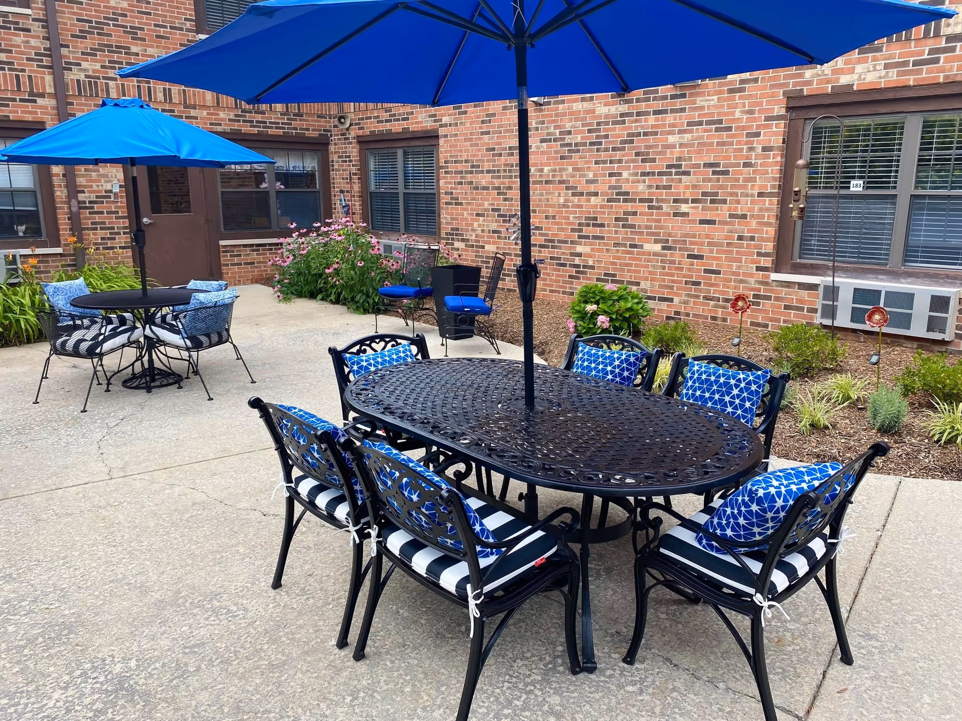 Outdoor patio area with black metal tables and chairs featuring blue and white cushions and blue umbrellas. The patio is adjacent to a brick building with windows and some plants and flowers along the edge.
