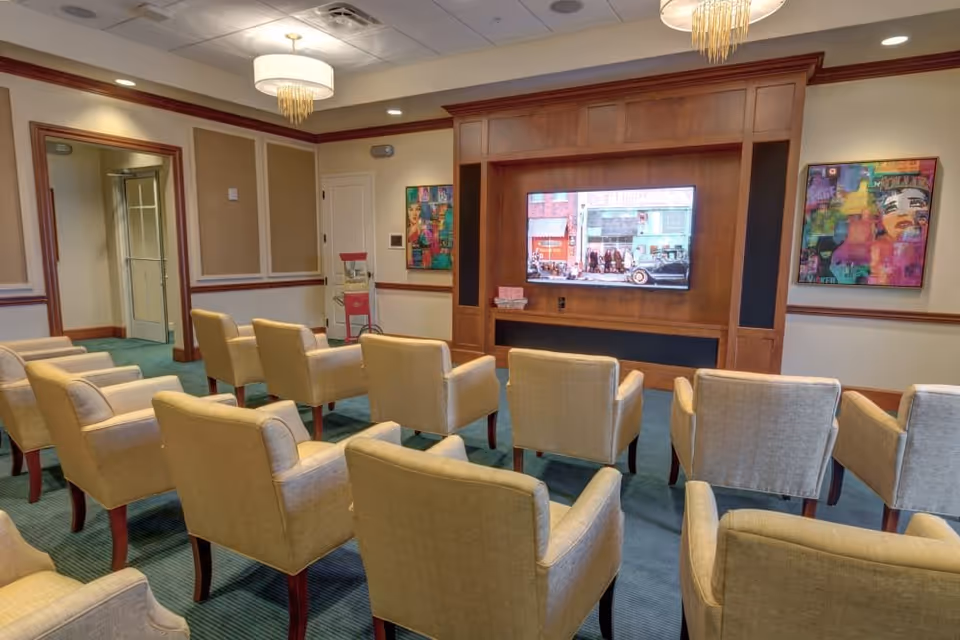 A small theater-style room with three rows of beige upholstered armchairs facing a large flat-screen TV mounted on a wooden entertainment center. The room has green carpet, cream-colored walls with wood trim, two colorful abstract paintings on the walls, and a popcorn machine in the corner. A modern chandelier hangs from the ceiling.