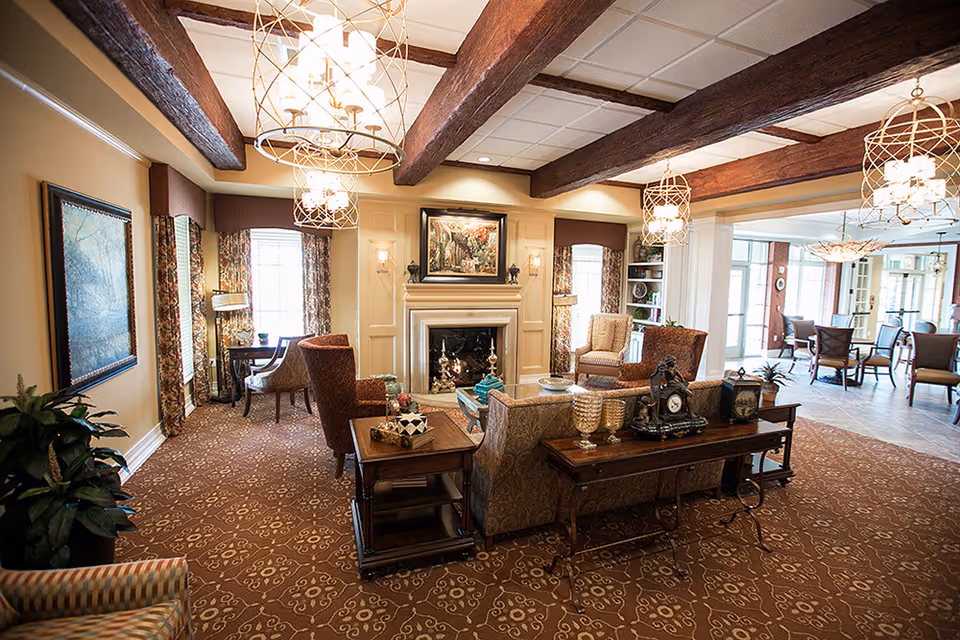 A cozy living room area in a senior living facility featuring a fireplace with a painting above it, comfortable upholstered chairs and sofas arranged around a wooden coffee table, decorative lamps hanging from a ceiling with exposed wooden beams, patterned carpet, and large windows with floral curtains allowing natural light to fill the space. In the background, there is a dining area with tables and chairs.