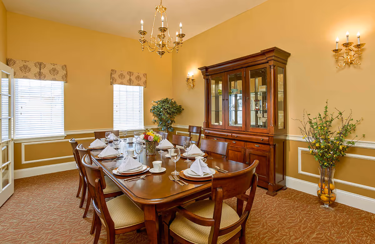 A formal dining room with a long wooden table set for eight people with white napkins, plates, cups, and glasses. The room features two windows with patterned valances, a wooden china cabinet filled with decorative items, a chandelier, wall sconces, and two large vases with plants in the corners.