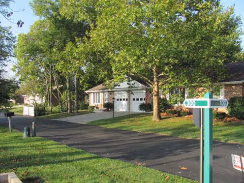 A tree-lined driveway with single-story homes and garages and a green address post in the foreground.