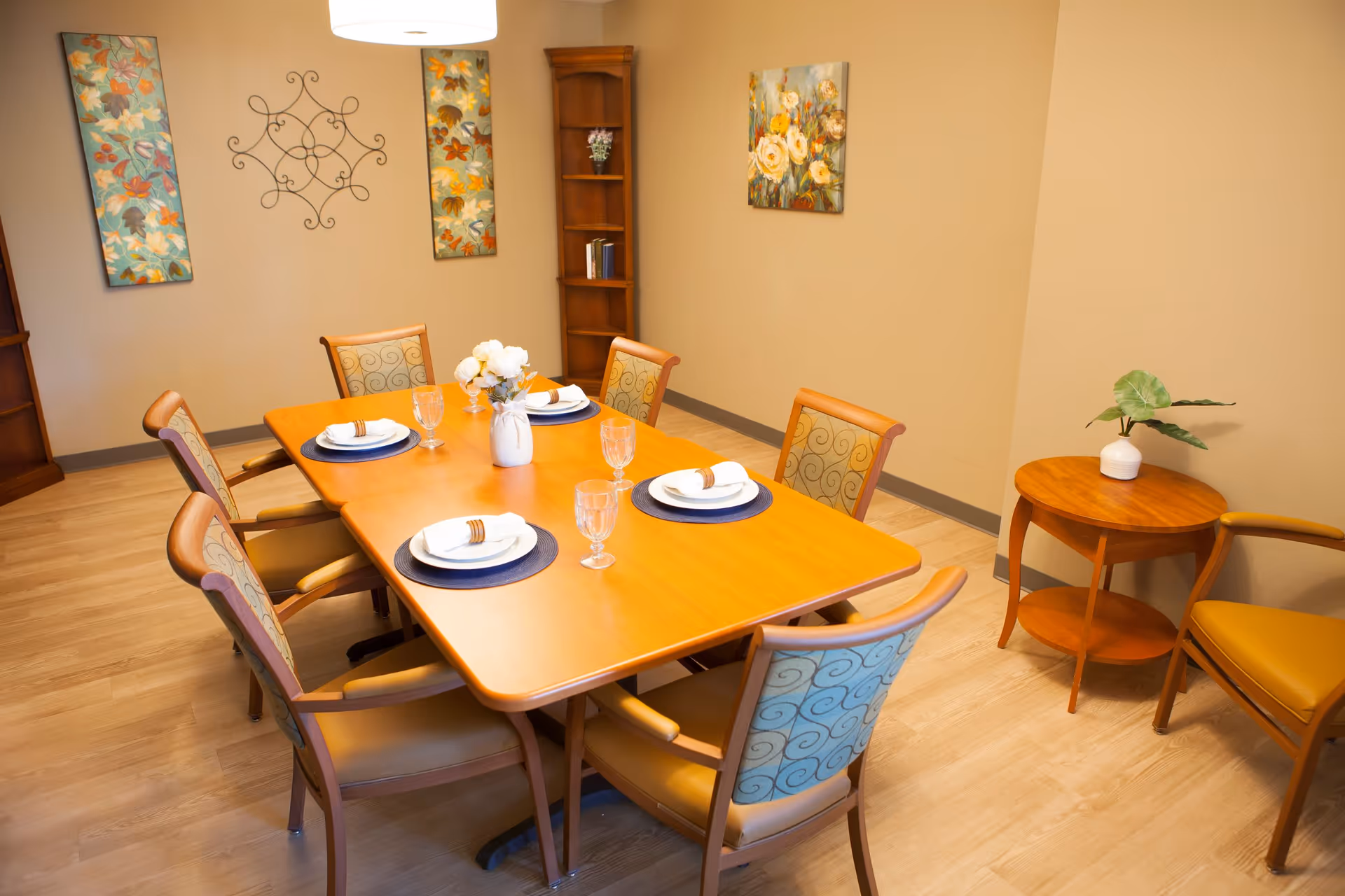 A dining room with a wooden table set for six people. Each place setting has a white plate with a napkin and a glass. The chairs around the table have patterned upholstery. The room has light-colored walls decorated with floral paintings and a metal wall art piece. There is a small round wooden side table with a plant in a white vase and an additional chair nearby. The floor is light wood.