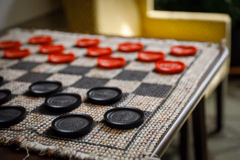 Close-up view of a checkers game board with black and red checkers pieces arranged on a woven fabric board, with a yellow chair in the background.