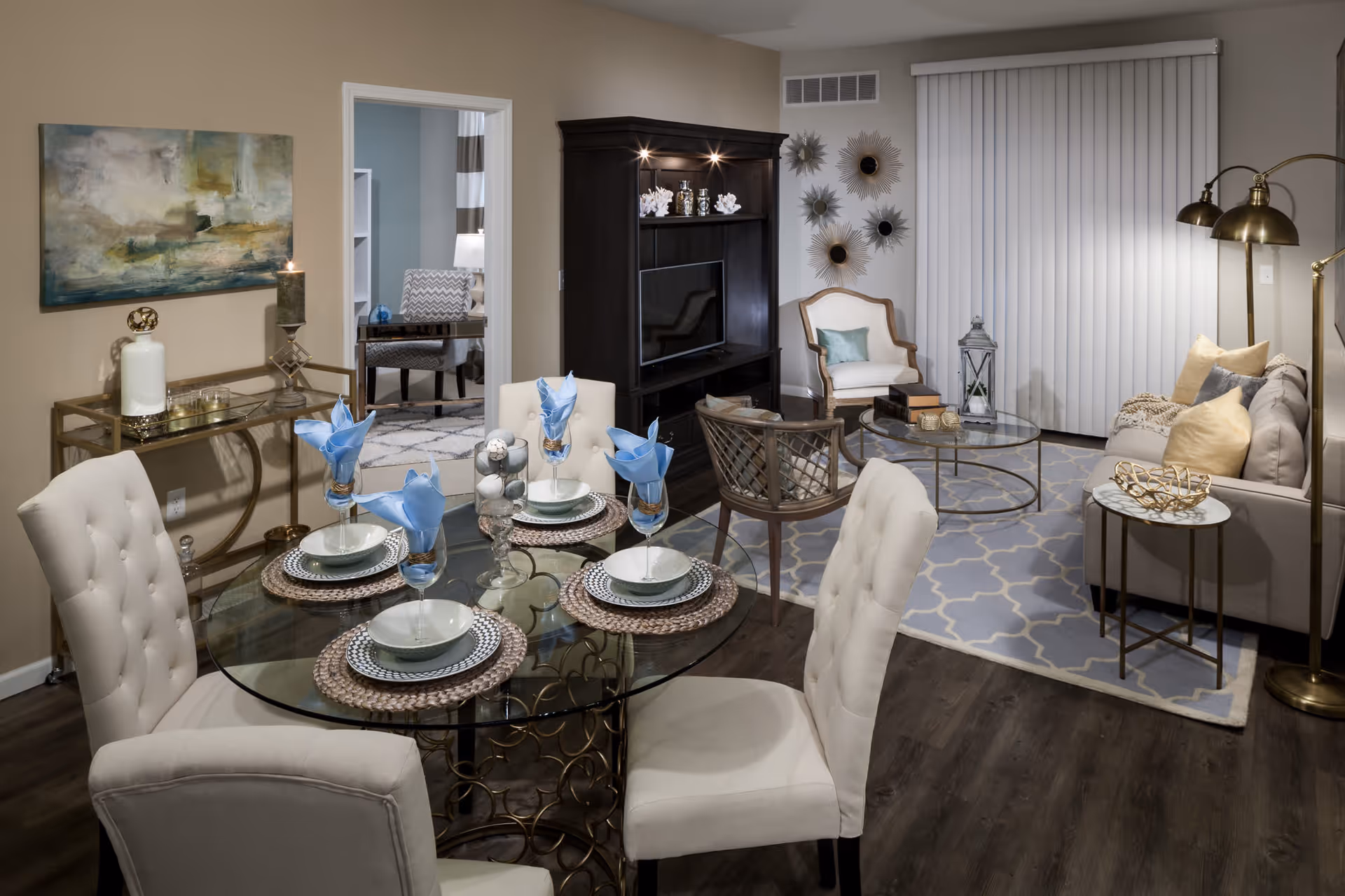 A cozy and elegantly decorated living and dining area in a senior living facility. The dining area features a round glass table set for four with white cushioned chairs and blue napkins. The living area includes a beige sofa with pillows, a glass coffee table, an armchair, a TV on a dark wooden stand, and decorative wall art. The room has wood flooring and vertical blinds covering a window or sliding door.