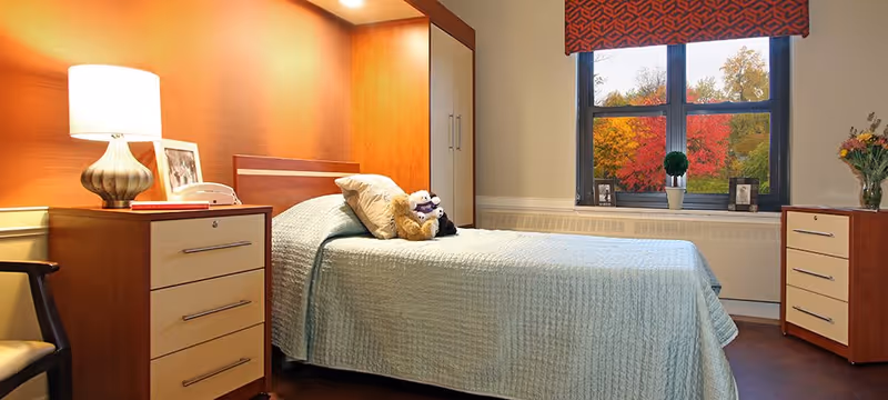 A neatly made single bed with a light blue quilt and stuffed animals on top, positioned against a wooden headboard and orange accent wall. To the left of the bed is a wooden nightstand with a lamp, a framed photo, and a telephone. A window with a red patterned valance shows colorful autumn trees outside. On the right side of the room is a wooden dresser with a vase of flowers on top. The room has a warm and cozy atmosphere.