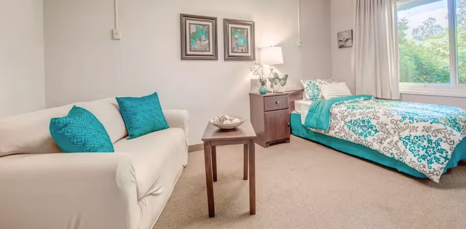A bright and cozy bedroom featuring a single bed with teal and white patterned bedding, a beige sofa with teal cushions, a wooden side table with decorative items, a lamp, two framed artworks on the wall, and a large window with white curtains letting in natural light.