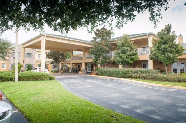 Exterior view of a senior living facility named Hunt Trace showing a two-story building with a covered entrance driveway, surrounded by green bushes and trees under a partly cloudy sky.