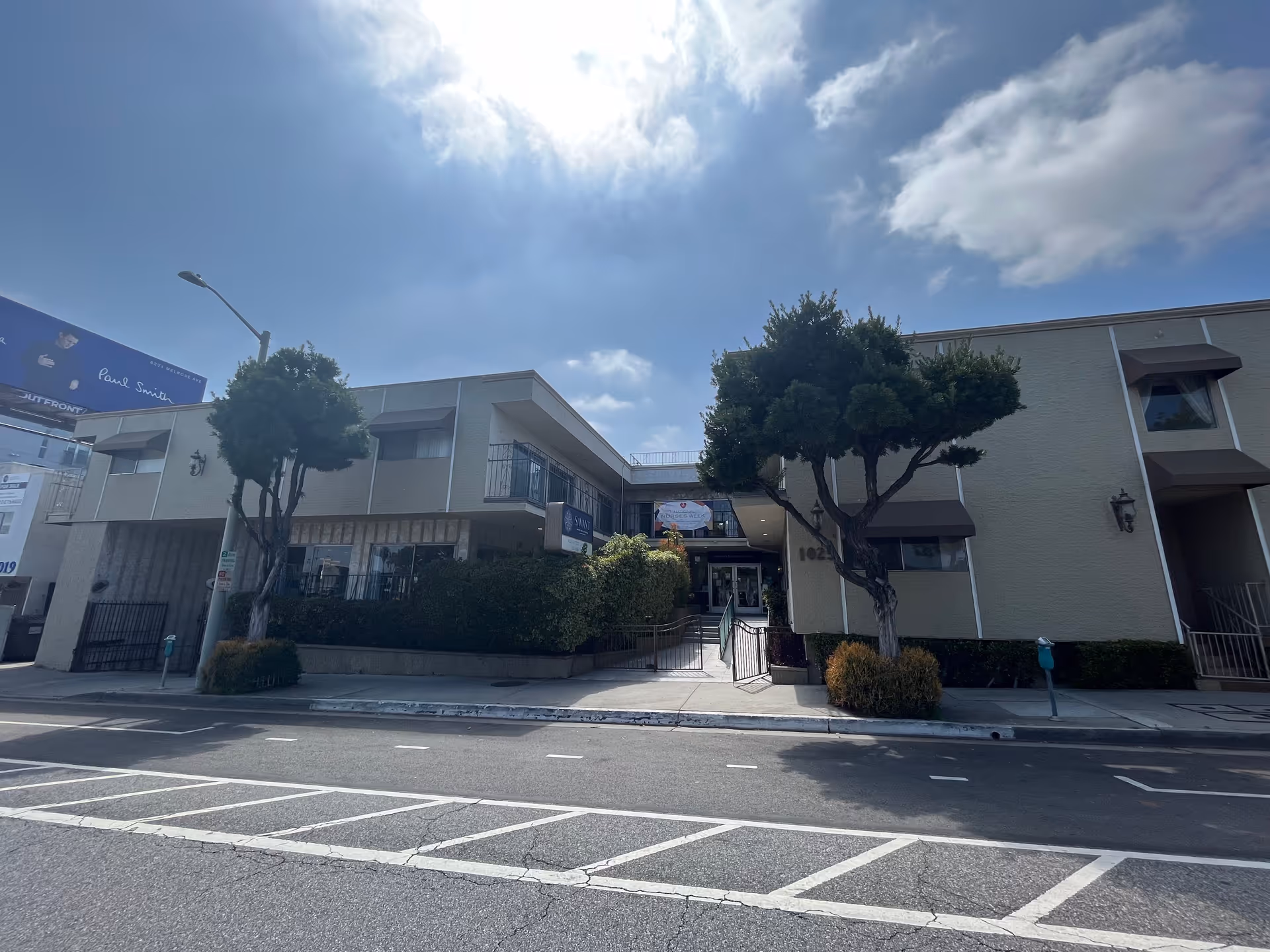 Exterior view of a two-story building with beige walls and brown awnings over the windows. There are two trees and some bushes in front of the building, and a ramp leading to the entrance. The sky is partly cloudy with the sun shining brightly.