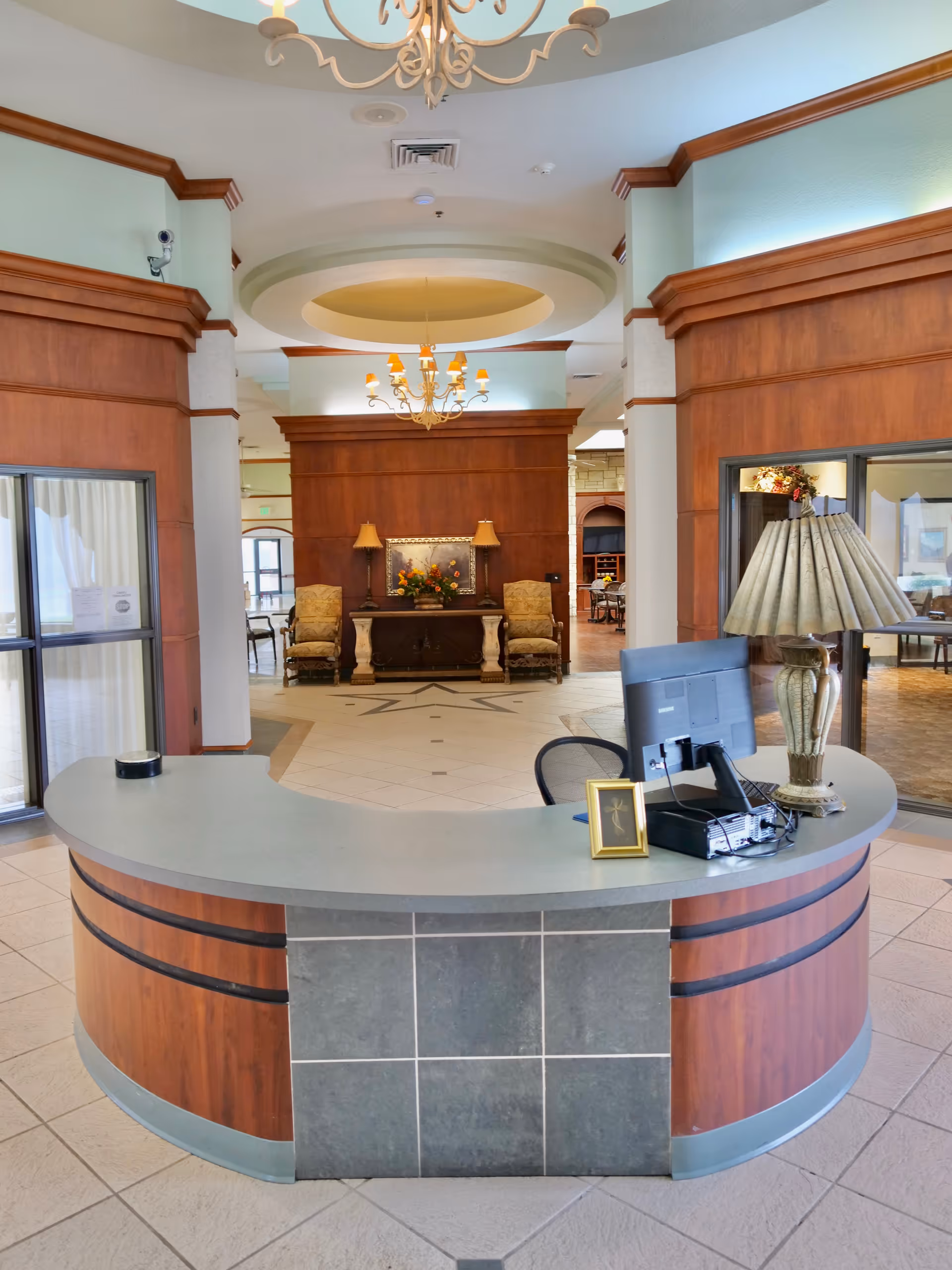 A nursing home reception desk in a spacious lobby with chandeliers, seating, and wood-paneled walls.