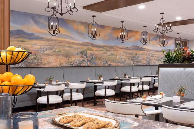 A dining room with multiple tables and white cushioned chairs arranged neatly. A long gray cushioned bench runs along the wall, which is decorated with a large mural depicting a desert landscape with mountains and vegetation. Several black metal pendant lights hang from the ceiling. A tiered fruit basket with oranges is visible in the foreground, along with a tray of cookies on a table.